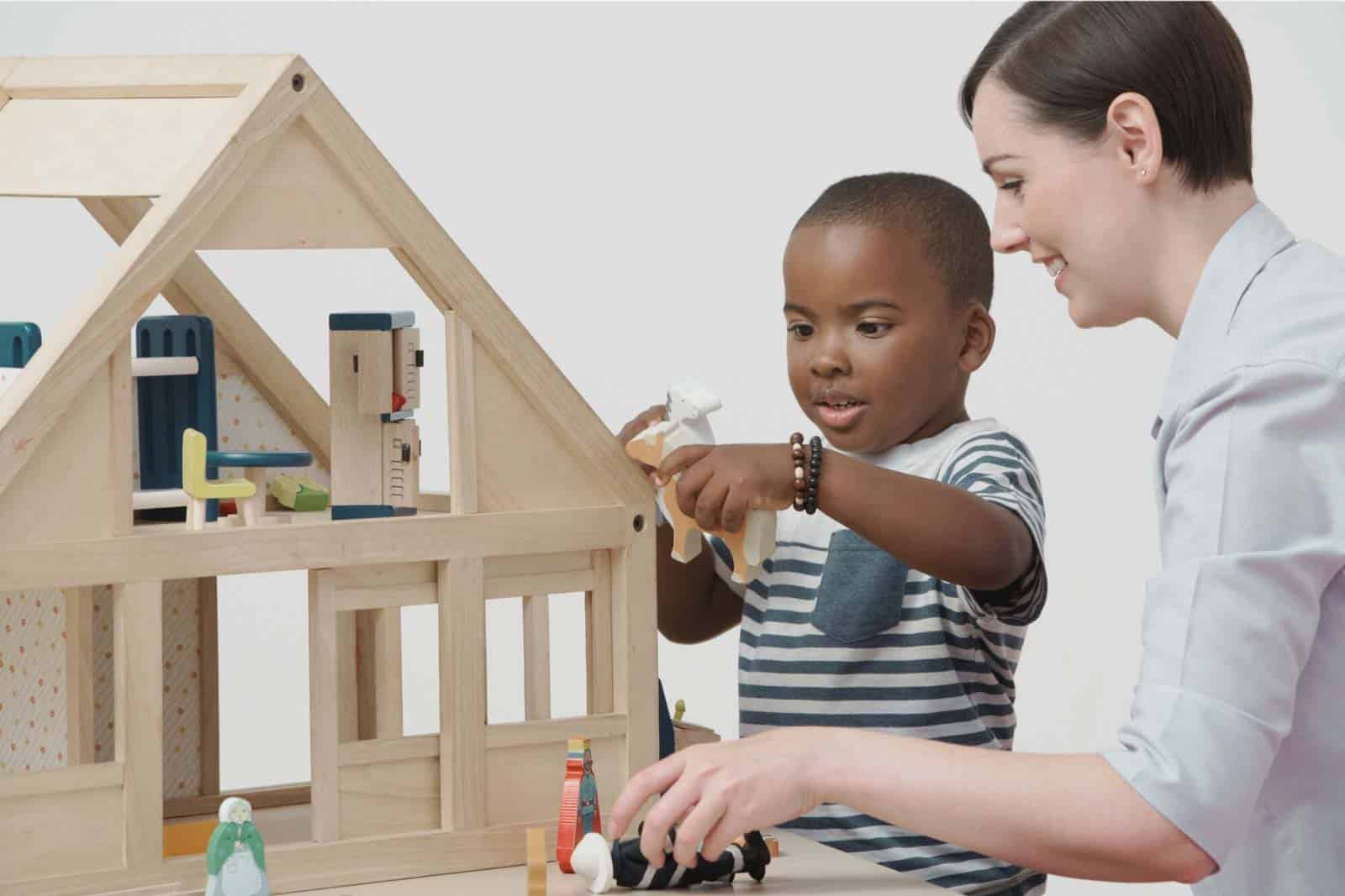 Child and caregiver playing with wooden dollhouse in childcare setting.