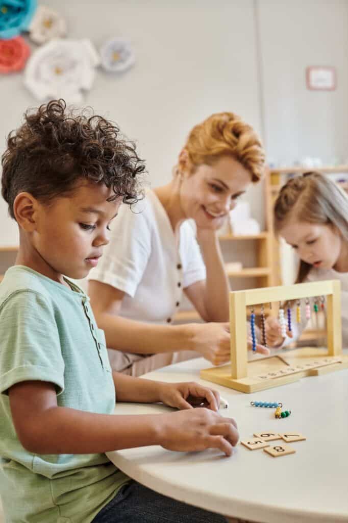 A teacher smiles at a table while a young girl uses a wooden bead counting frame and a young boy focuses on wooden number tiles in a bright classroom.